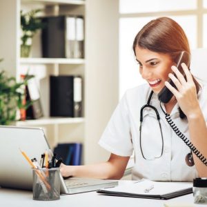Beautiful young smiling female doctor sitting in her consulting room using phone and working on laptop.